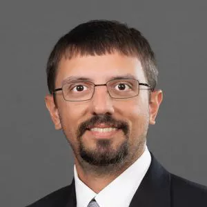 Donnie Smith, a white man with stright dark hair and a goatee, wearing rectangular wire-frame glasses, against a dark gray backdrop