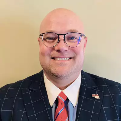 Jason Culler, a White man, bald with round thin-framed glasses, smiling, wearing a navy checkered sports jacket with a white dress shirt, and red and light blue stripped tie, with an Oklahoma pin on the lapel.