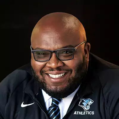 Ryan Doggett, a Black man with a short-cut beard, smiling, wearing a navy-colored Nike quarter zip over a white dress shirt with a stripped tie of navy and sky blue